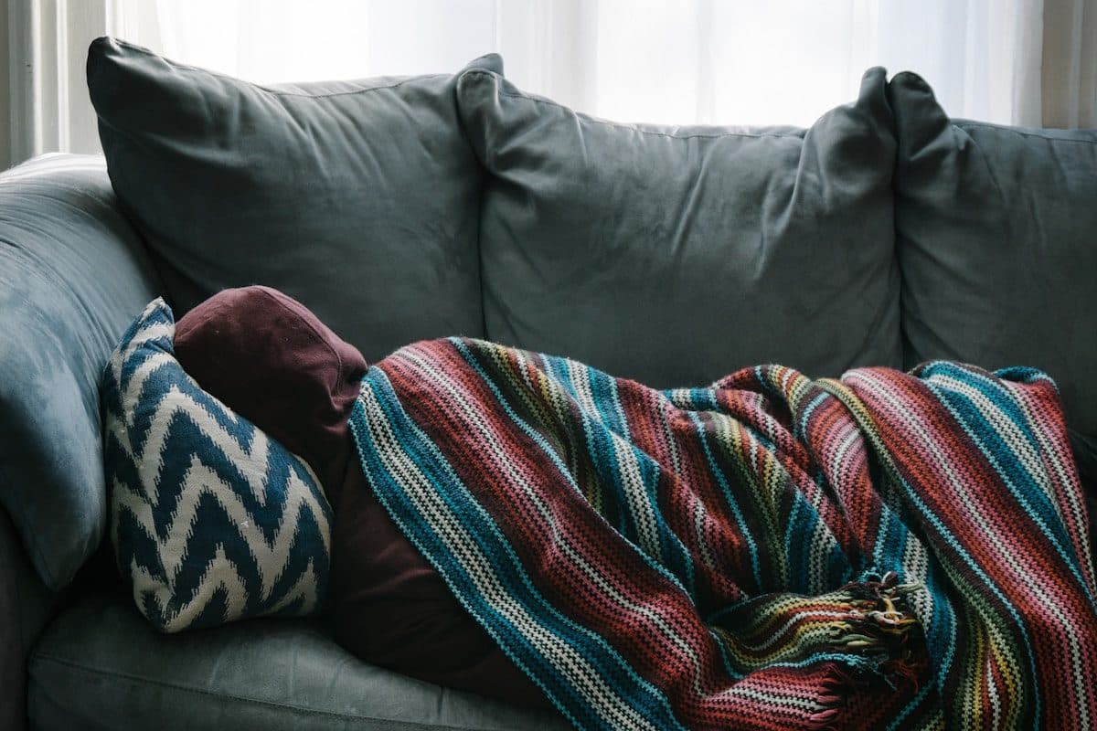 Person resting under a striped crochet blanket on a grey sofa by a window